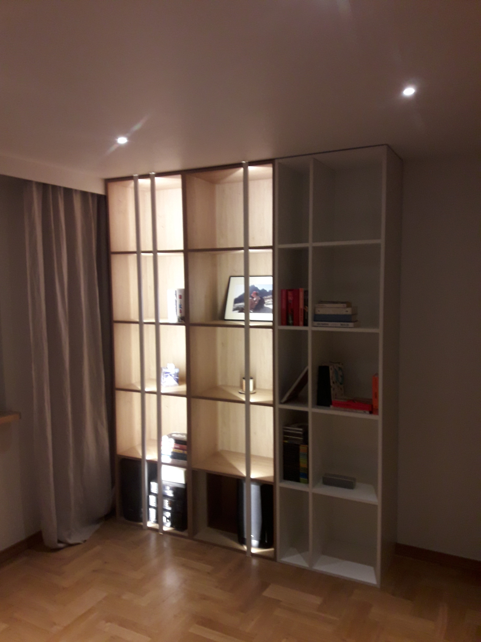 Regał modułowy z otwartymi półkami, część drewniana z oświetleniem wewnętrznym, część biała, ustawiony w rogu pokoju z parkietem i zasłoniętym oknem.