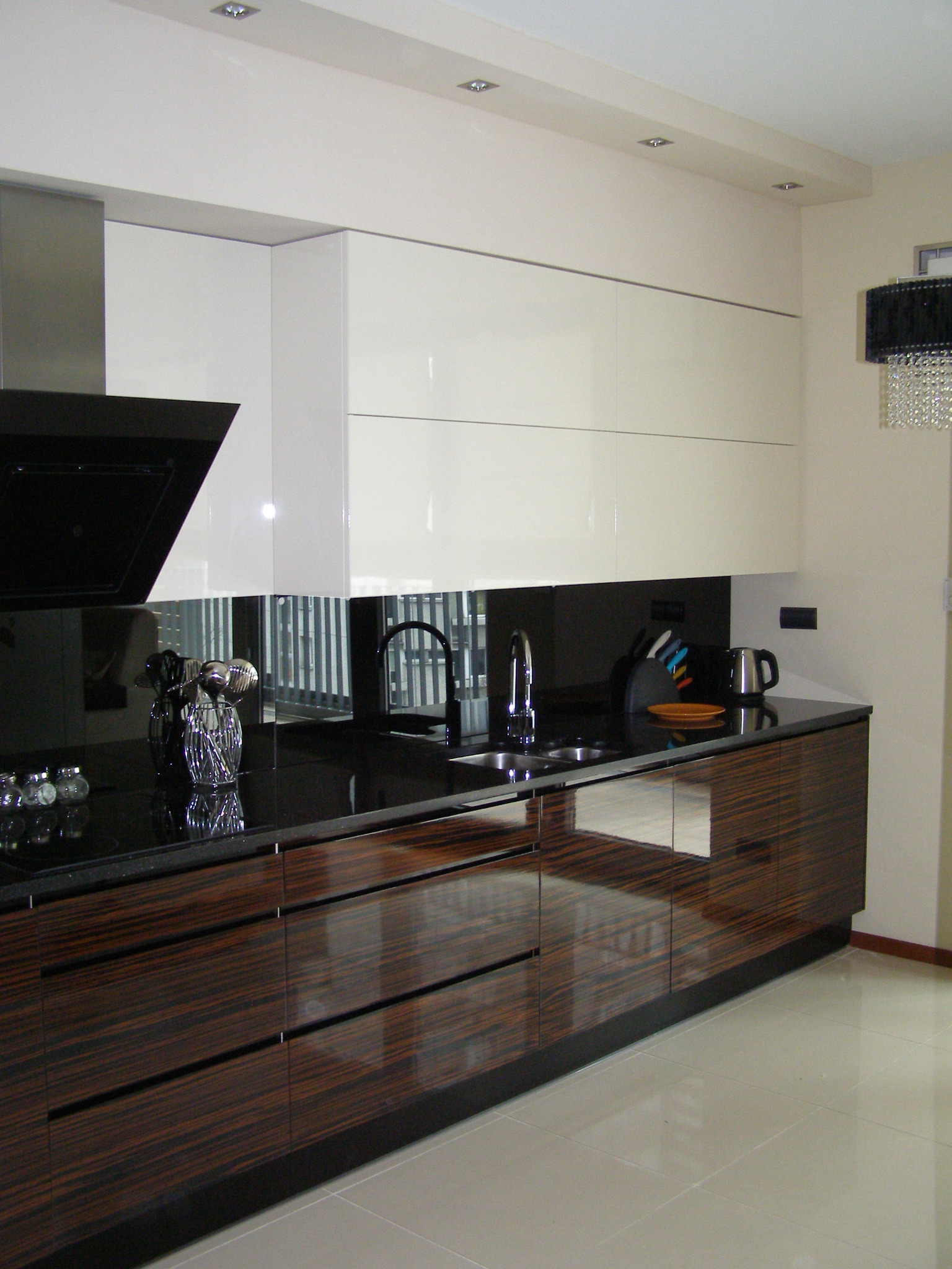 Nowoczesna kuchnia z ciemnym blatem i dolnymi szafkami z egzotycznego drewna, górne szafki w kolorze złamanej bieli, czarny panel nad blatem, oświetlenie punktowe i dekoracyjny żyrandol.