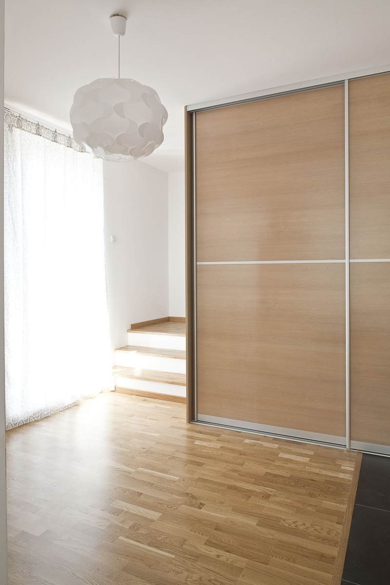 Jasne wnętrze z drewnianą podłogą i przesuwną szafą z jasnego drewna, widoczne schody w tle i designerska lampa wisząca.