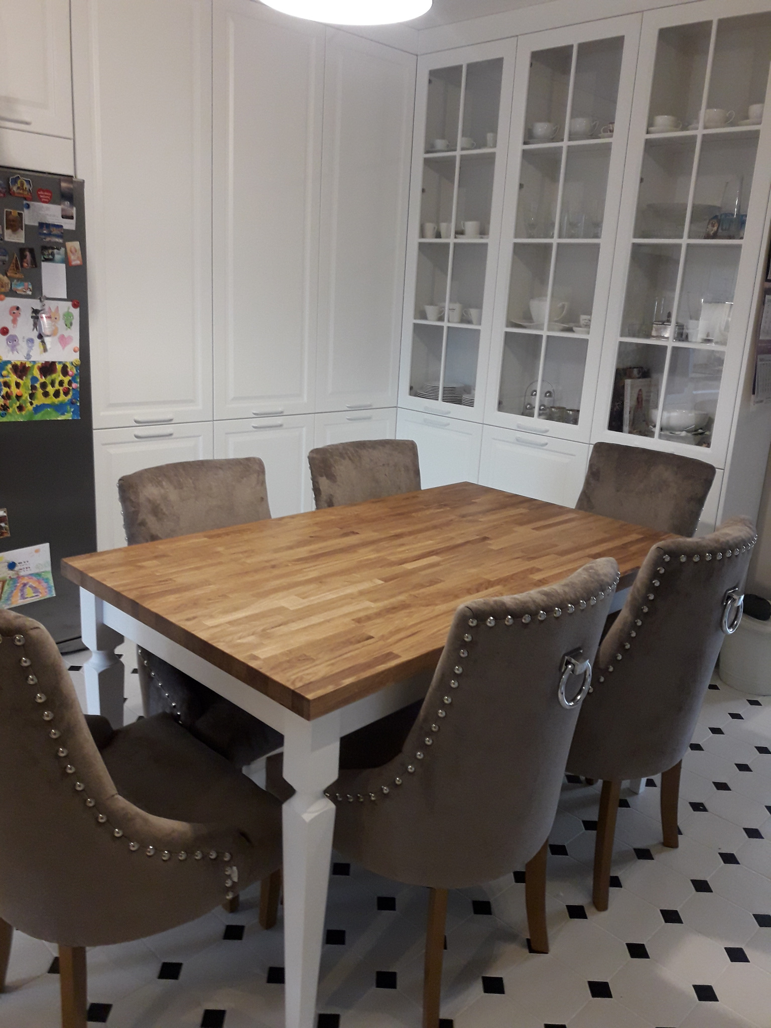 Jasna kuchnia z drewnianym blatem stołu i krzesłami obitymi szarą tkaniną z ozdobnymi nitami, widoczne białe szafki kuchenne z przeszklonymi witrynami.