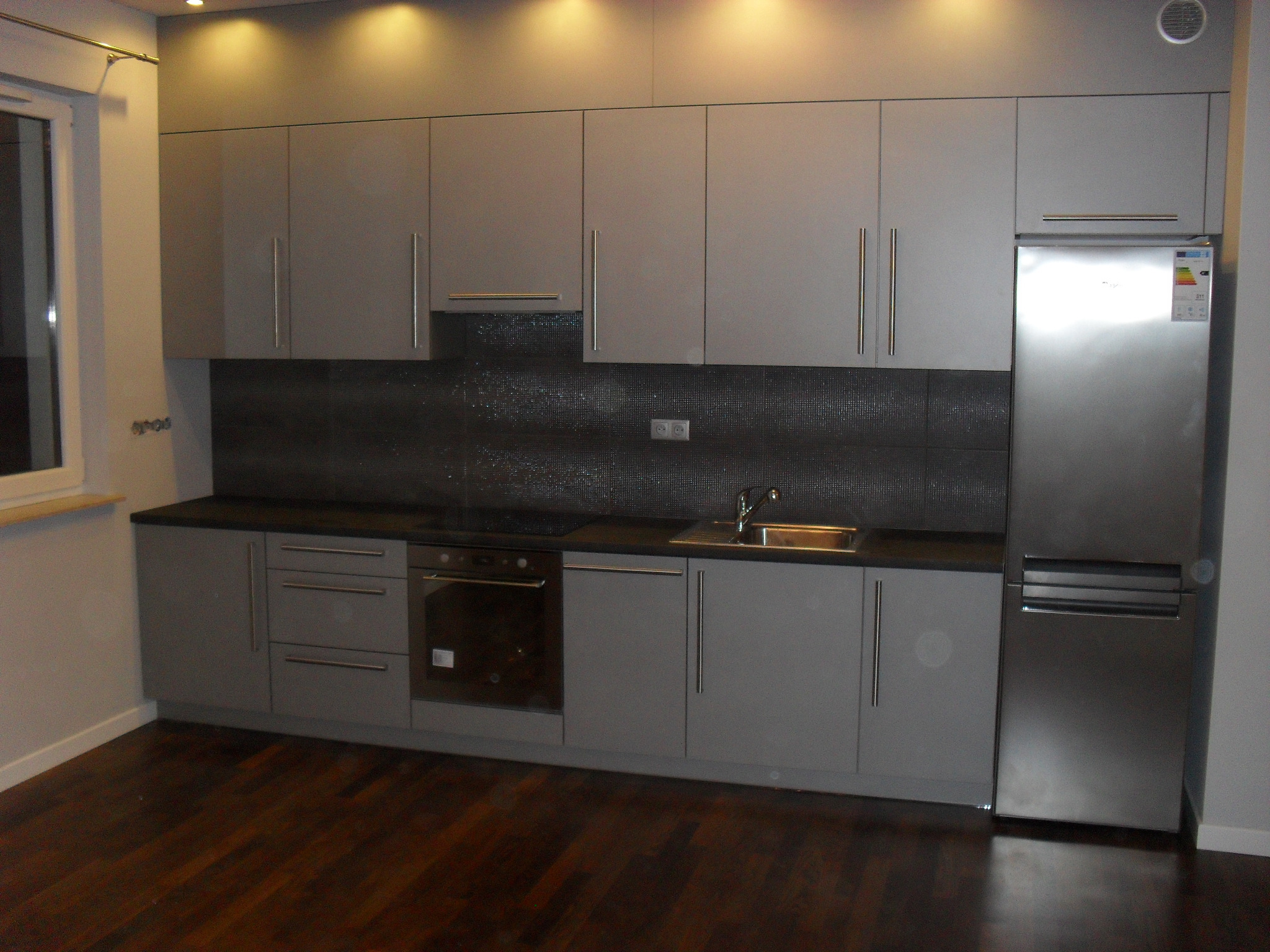 Nowoczesna kuchnia w odcieniach szarości z ciemnym blatem i mozaikową ścianą, oświetlona punktowymi lampami sufitowymi, z widoczną stalową lodówką.