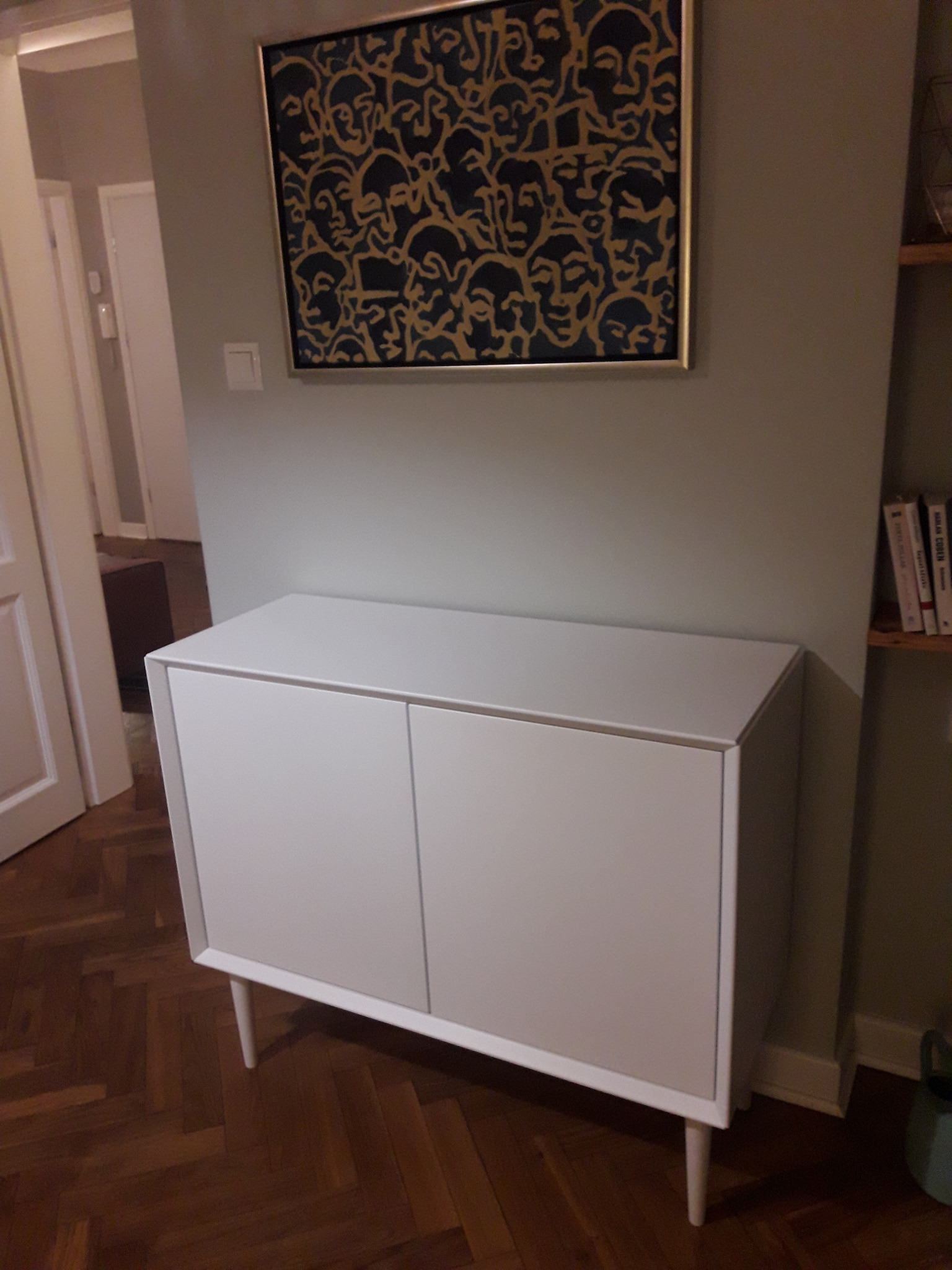 Biała szafka z dwiema drzwiczkami na drewnianych nóżkach, ustawiona na parkiecie w jodełkę, z obrazem przedstawiającym stylizowane twarze zawieszonym nad nią.