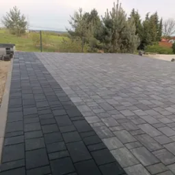 Ułożona nawierzchnia z kostki brukowej w dw&oacute;ch odcieniach szarości, przechodząca w tle w zielony krajobraz z drzewami i ogrodzeniem z siatki.