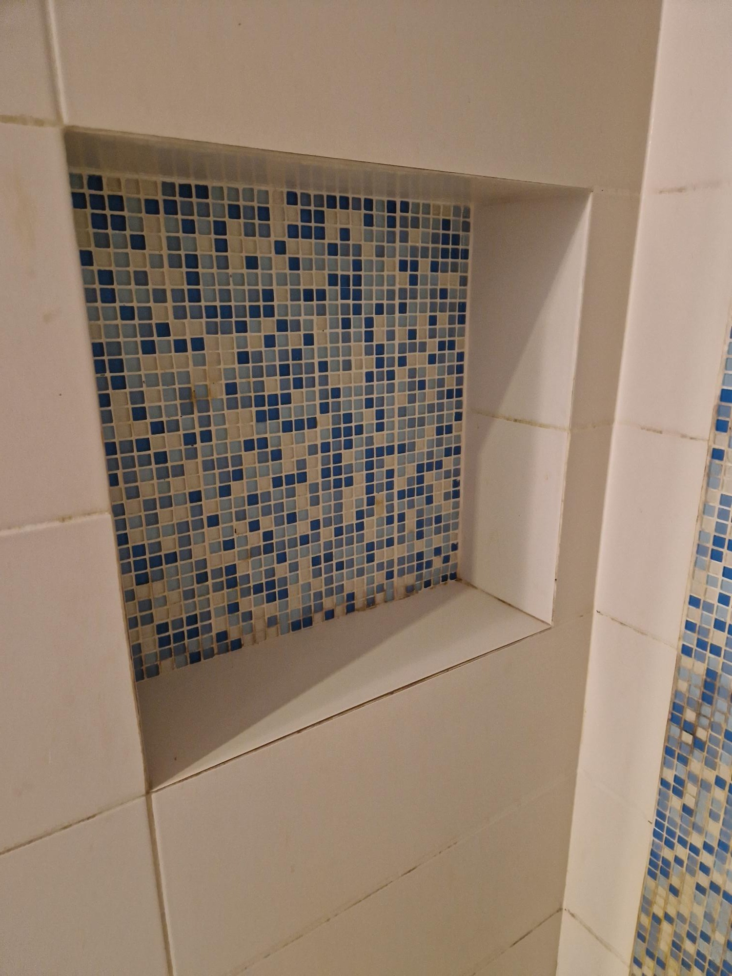 Wnęka w ścianie wyłożona mozaiką z niebiesko-białych płytek, otoczona białymi płytkami ceramicznymi. Widoczne fugi. Świeżo położone płytki.