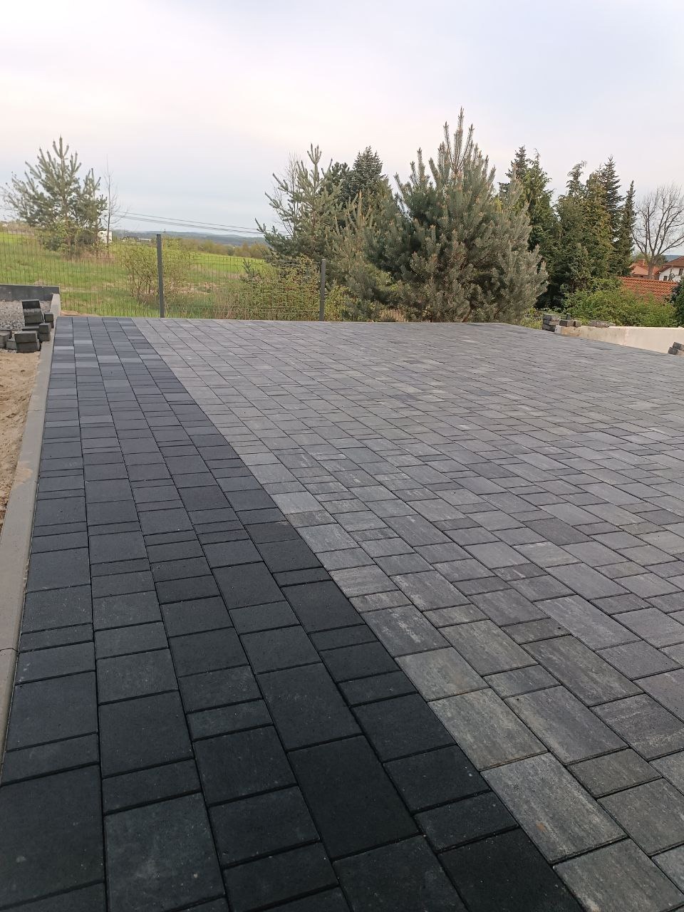 Ułożona nawierzchnia z kostki brukowej w dwóch odcieniach szarości, przechodząca w tle w zielony krajobraz z drzewami i ogrodzeniem z siatki.