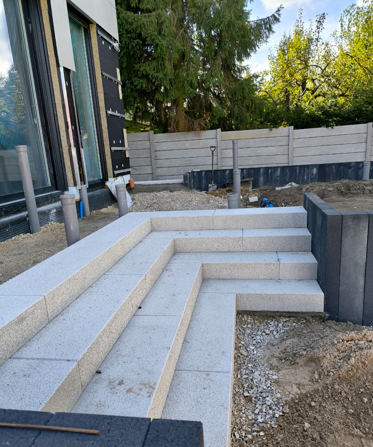 Schody zewnętrzne z jasnej kostki granitowej prowadzące do nowoczesnego domu, w tle widoczny betonowy płot i zieleń.