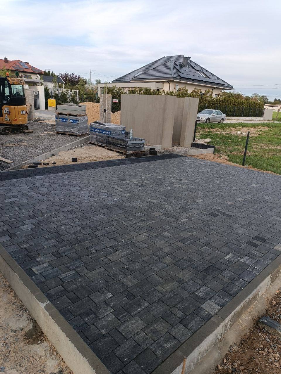 Układanie ciemnoszarej kostki brukowej na podjeździe, widoczny mały żółty traktor i palety z kostką w tle, dom jednorodzinny w oddali.