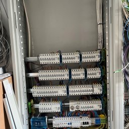 MG ENERGY Instalacje elektryczne - Rozdzielnia elektryczna w trakcie montażu, widoczne zabezpieczenia nadprądowe, przewody oznaczone etykietami, szyna DIN, gniazdo elektryczne i elementy instalacji w metalowej obudowie.