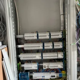 Rozdzielnia elektryczna w trakcie montażu, widoczne zabezpieczenia nadprądowe, przewody oznaczone etykietami, szyna DIN, gniazdo elektryczne i elementy instalacji w metalowej obudowie.