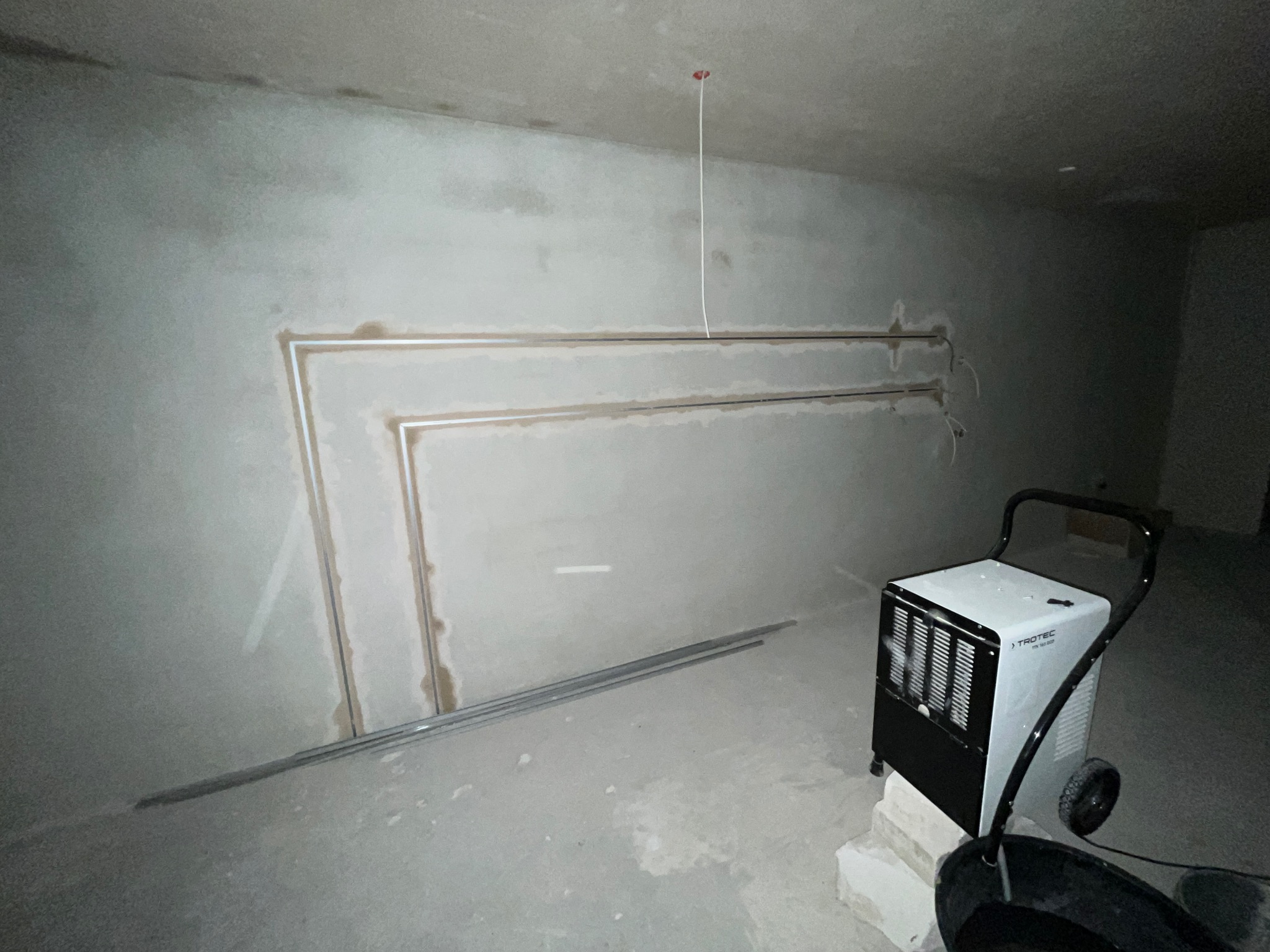 Surowa ściana z przygotowanymi bruzdami pod przewody, widoczny przewód zwisający z sufitu i osuszacz powietrza TROTEC na cegłach.