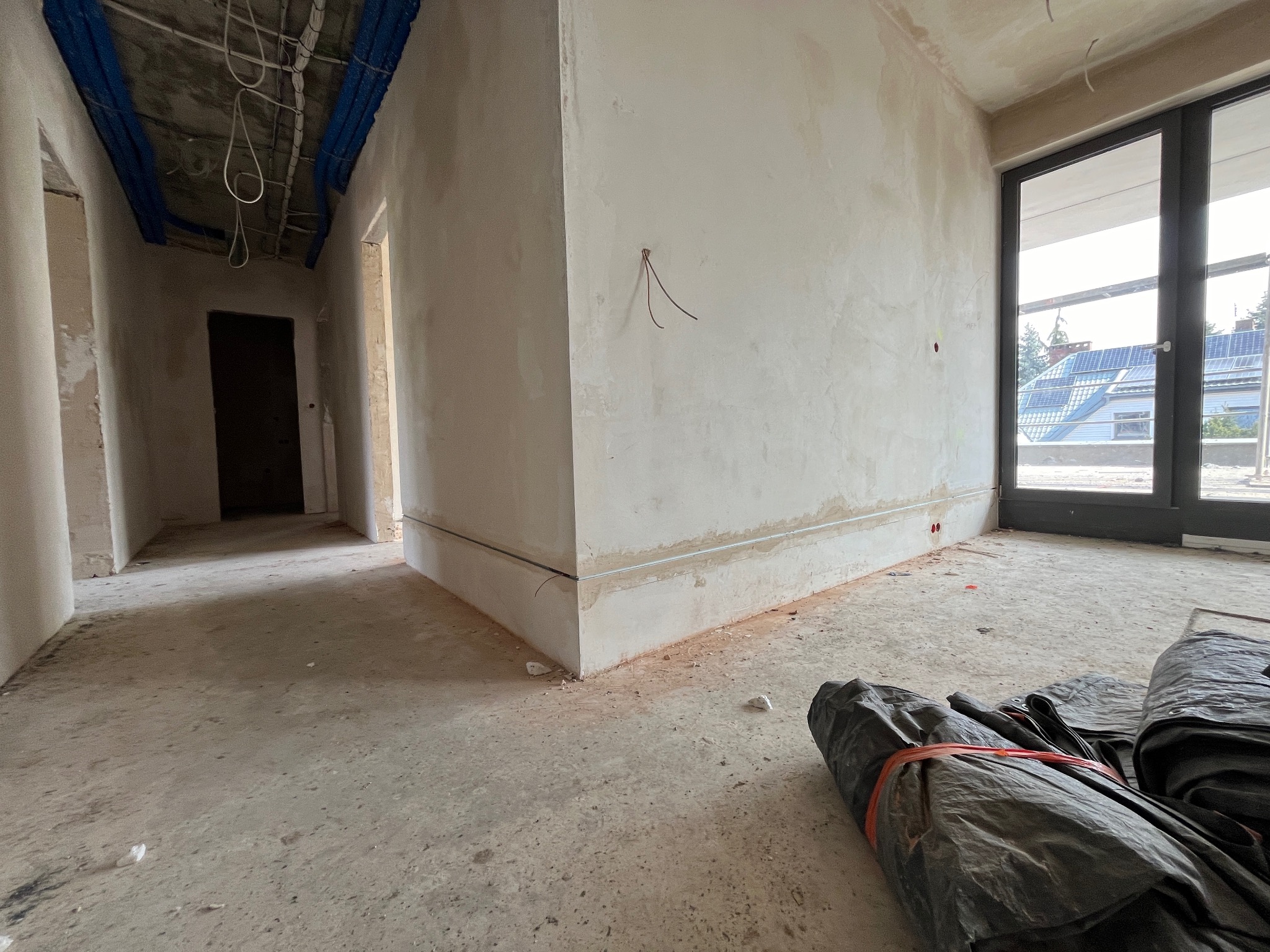 Surowe wnętrze w trakcie wykańczania: widoczne kable wystające ze ściany, rury instalacyjne pod sufitem, betonowa podłoga i okno z widokiem na panele słoneczne na dachu sąsiedniego budynku.