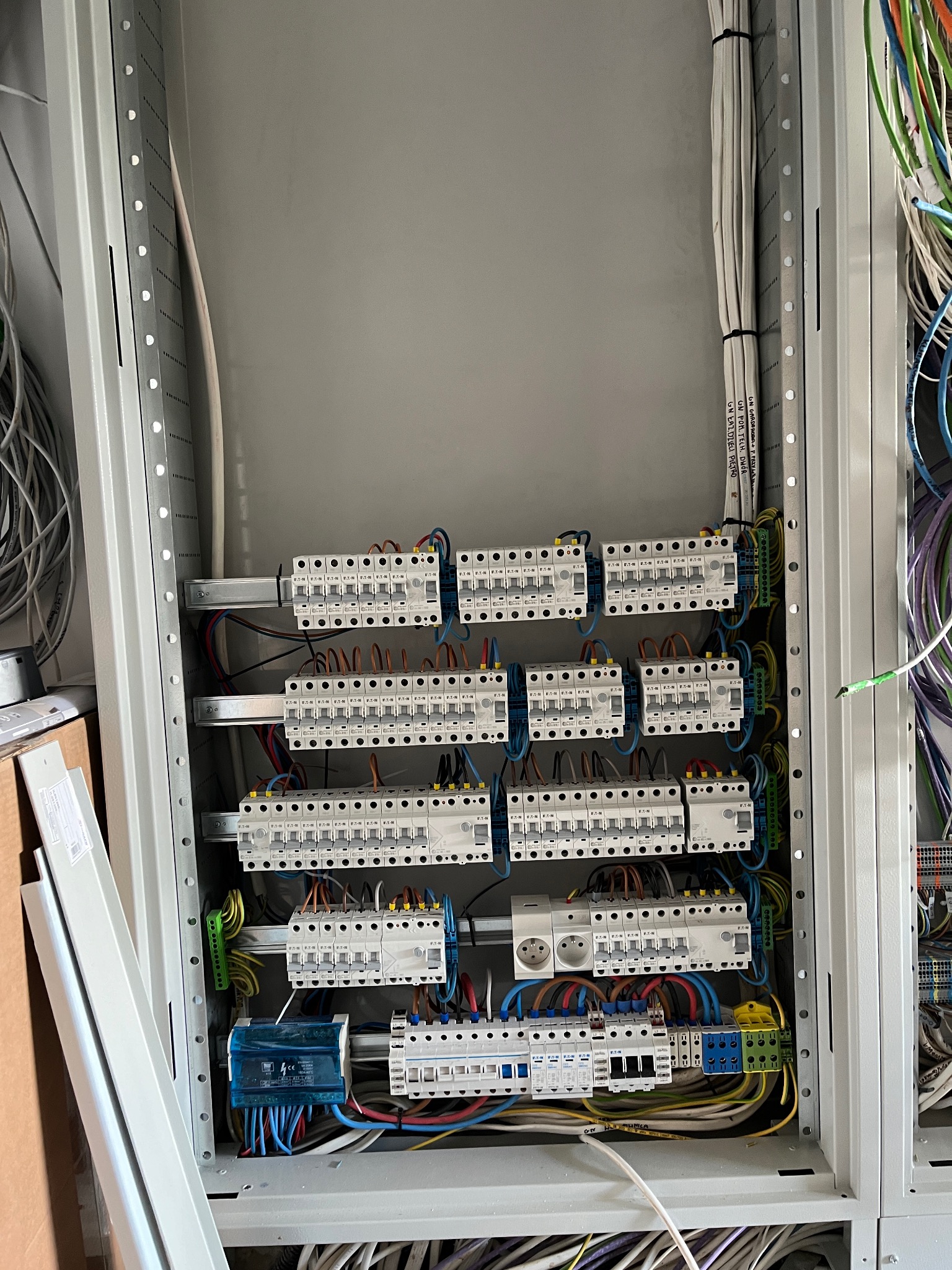 Rozdzielnia elektryczna w trakcie montażu, widoczne zabezpieczenia nadprądowe, przewody oznaczone etykietami, szyna DIN, gniazdo elektryczne i elementy instalacji w metalowej obudowie.