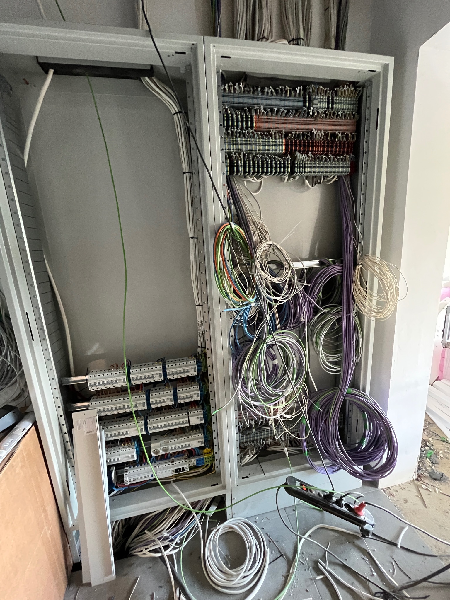 Dwa otwarte panele elektryczne w trakcie montażu okablowania. Widoczne liczne przewody w różnych kolorach, listwy zaciskowe i elementy montażowe. Na podłodze leżą dodatkowe kable i listwa zasilająca.