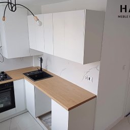 Mateusz Wendel - Nowoczesna, minimalistyczna kuchnia z białymi szafkami i blatem w kolorze jasnego drewna, czarny zlew i płyta grzewcza, marmurowe płytki na ścianie, widoczne logo firmy meblarskiej w prawym górnym...