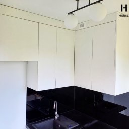 Mateusz Wendel - Nowoczesna kuchnia z białymi szafkami górnymi i czarnym blatem, oświetlona lampą z dwoma kulistymi kloszami. Widoczne logo 'HALI MEBLE NA WYMIAR' w prawym górnym rogu.