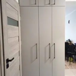 Jasnoszara szafa z czterema drzwiami i długimi, czarnymi uchwytami, obok drewniane drzwi z przeszkleniem, widoczne logo firmy na szafie.