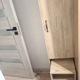 Jasna szafa z drewnopodobnym dekorem, wbudowana obok drzwi z czarną klamką, z otwartą półką na dole i logo firmy HALI u góry.