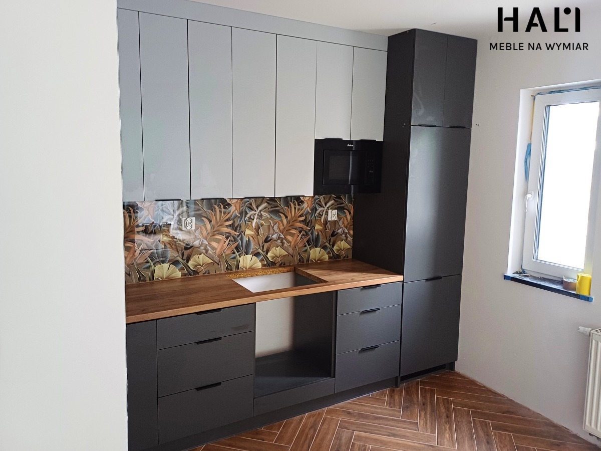 Nowoczesna kuchnia z szarymi szafkami górnymi i grafitowymi dolnymi, drewniany blat, wzorzysta ściana nad blatem, parkiet w jodełkę. Logo firmy widoczne w prawym górnym rogu.