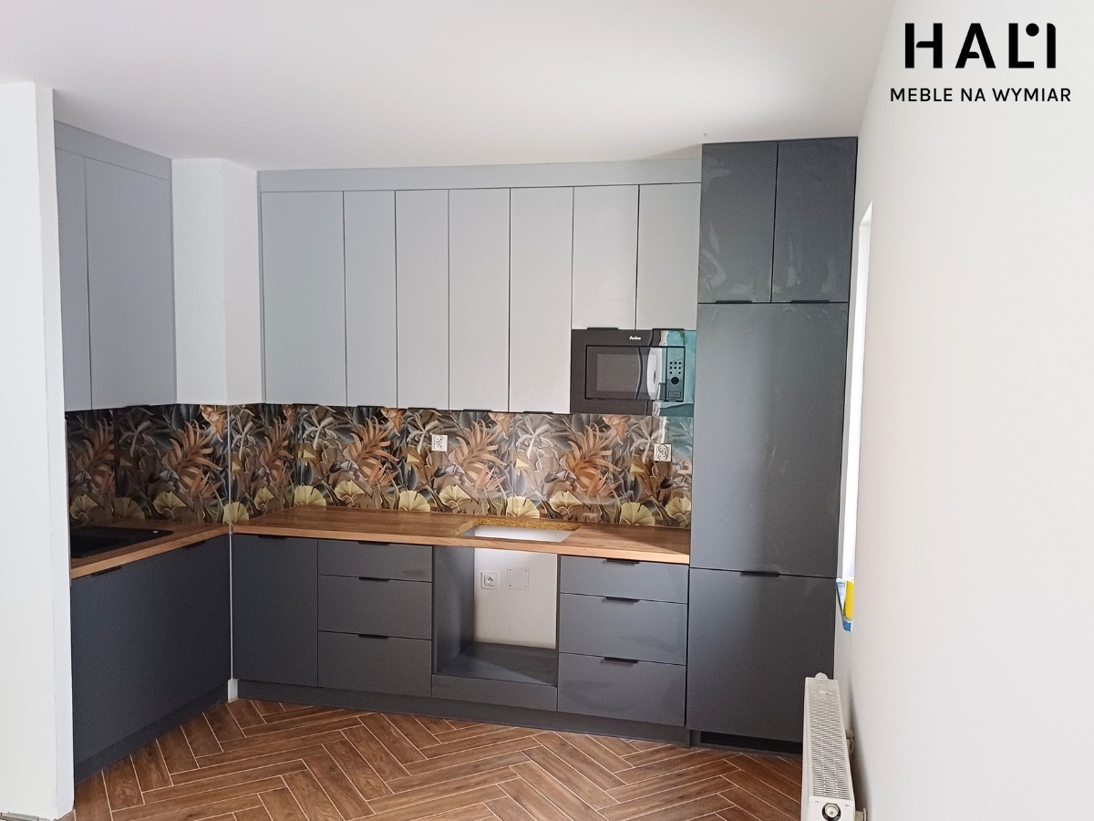 Szaro-biała zabudowa kuchenna z drewnianym blatem i wzorzystą ścianą nad blatem, uzupełniona o mikrofalówkę Amica, na podłodze parkiet w jodełkę.