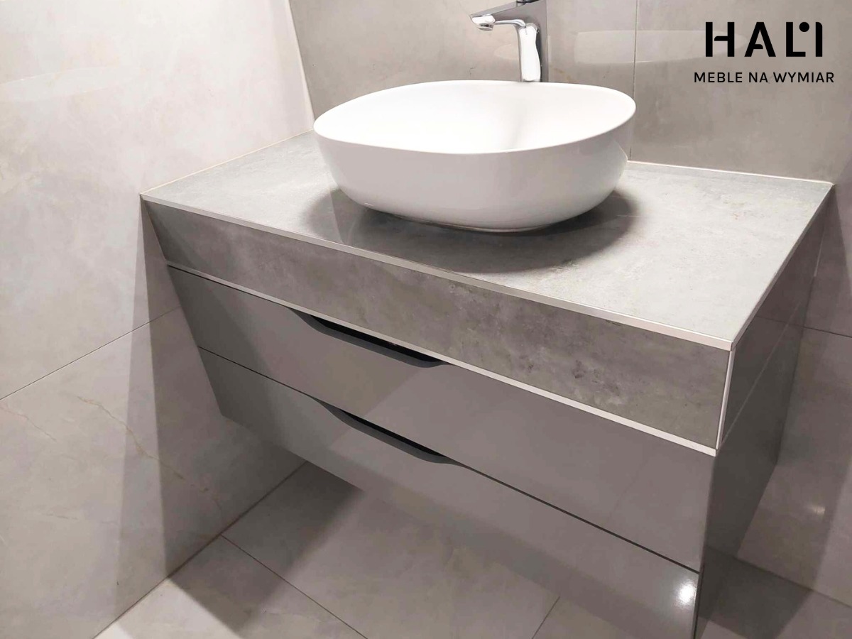 Szara szafka łazienkowa z trzema szufladami, blat z efektem betonu, umywalka nablatowa owalna, minimalistyczna armatura, widoczne logo firmy meblowej w górnym rogu.