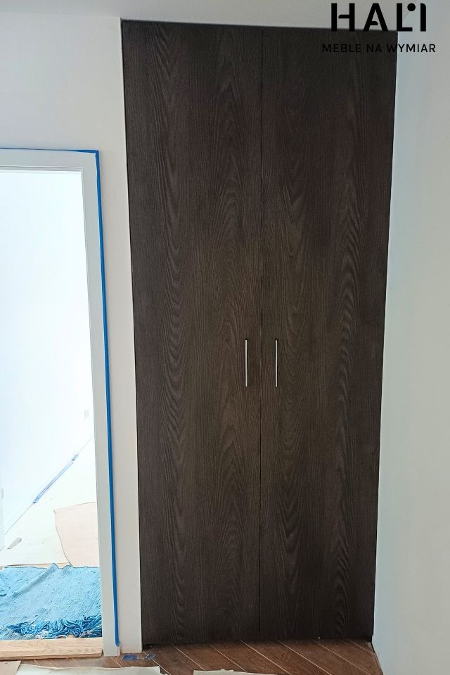 Ciemna szafa wnękowa z dwoma drzwiami i minimalistycznymi uchwytami, widoczne zabezpieczenia podłogi i futryny drzwi, logo firmy HALI na górze.