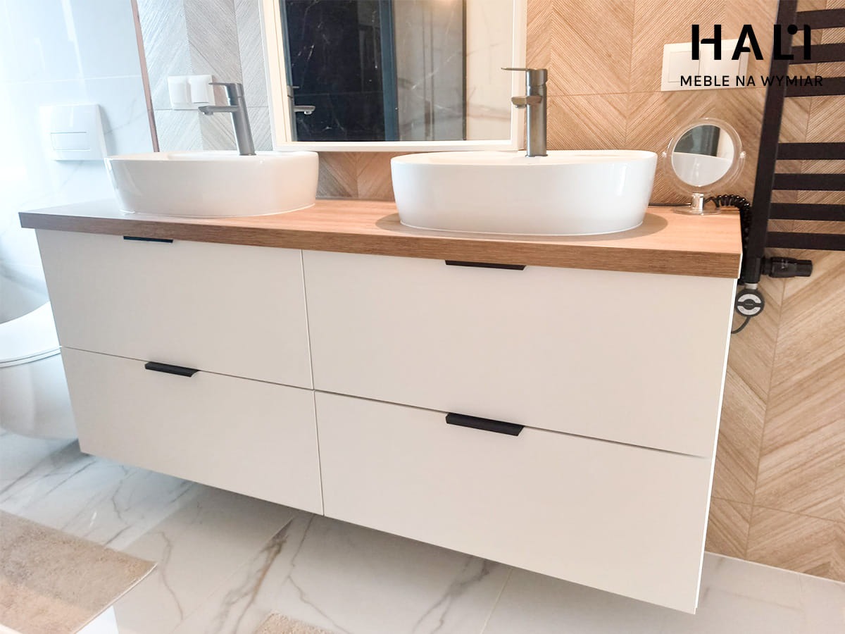 Łazienkowa szafka pod umywalki nablatowe z białymi frontami i drewnianym blatem, czarne uchwyty, nowoczesny grzejnik drabinkowy w tle, logo firmy na ścianie.