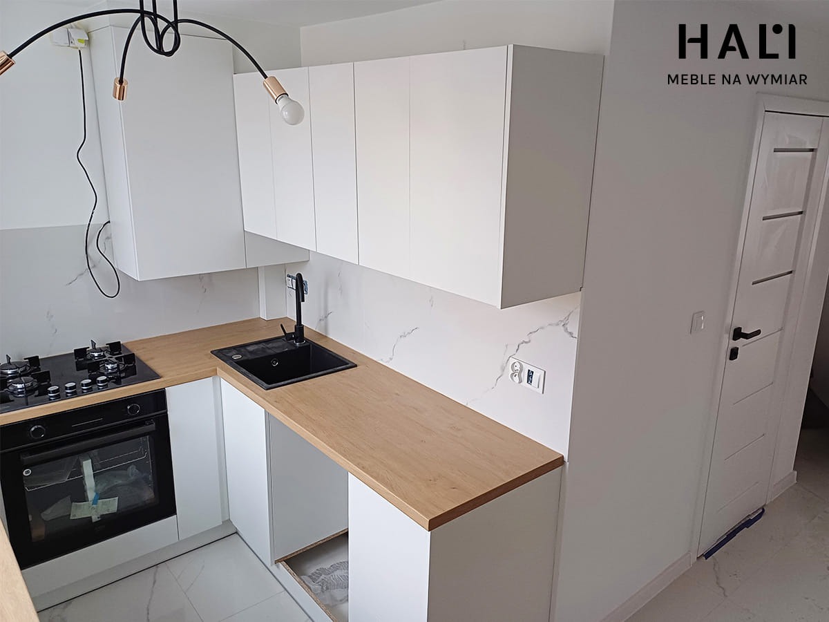 Nowoczesna, minimalistyczna kuchnia z białymi szafkami i blatem w kolorze jasnego drewna, czarny zlew i płyta grzewcza, marmurowe płytki na ścianie, widoczne logo firmy meblarskiej w prawym górnym...