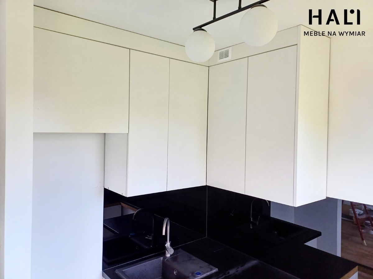 Nowoczesna kuchnia z białymi szafkami górnymi i czarnym blatem, oświetlona lampą z dwoma kulistymi kloszami. Widoczne logo 'HALI MEBLE NA WYMIAR' w prawym górnym rogu.