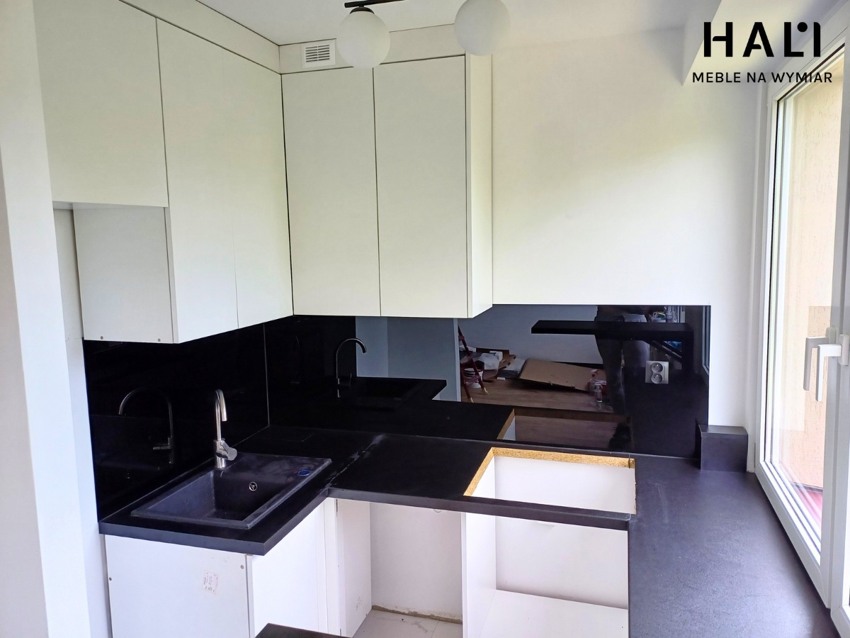 Nowoczesna kuchnia z białymi szafkami górnymi i czarnym blatem oraz szkłem na ścianie, widoczny montaż zlewu i niedokończona zabudowa dolna, logo firmy w prawym górnym rogu.