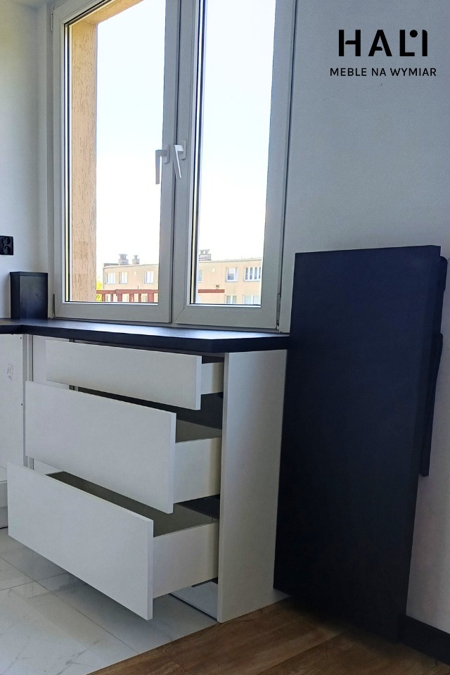 Biała szafka z trzema wysuwanymi szufladami pod oknem, czarny blat, widoczny budynek za oknem, obok pionowa czarna płyta.