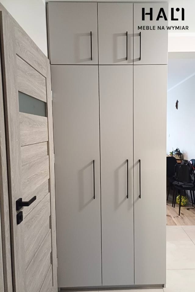 Jasnoszara szafa z czterema drzwiami i długimi, czarnymi uchwytami, obok drewniane drzwi z przeszkleniem, widoczne logo firmy na szafie.