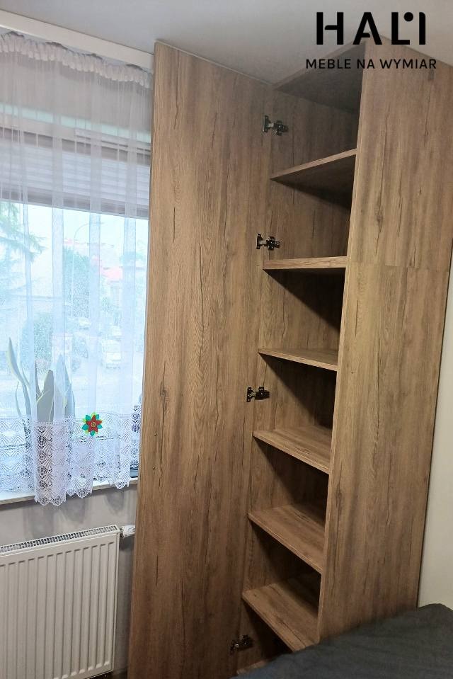 Szafa na wymiar z drewnopodobnym fornirem, otwarte drzwi ukazują puste półki, widoczny grzejnik pod oknem z firanką.