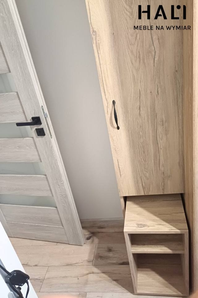 Jasna szafa z drewnopodobnym dekorem, wbudowana obok drzwi z czarną klamką, z otwartą półką na dole i logo firmy HALI u góry.
