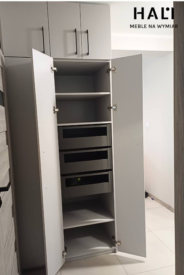 Szara szafa z otwartymi drzwiami, widoczne półki i trzy szuflady z ciemnoszarymi frontami, czarne uchwyty, logo firmy meblowej w prawym górnym rogu.