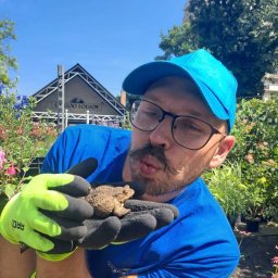 Łukasz Leśniewicz Ogrodnictwo - Mężczyzna w niebieskiej koszulce i czapce, z wąsami i okularach, trzyma w rękawicach ropuchę na tle szkółki roślin.