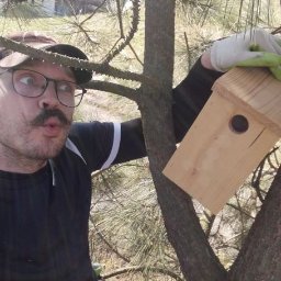 Łukasz Leśniewicz Ogrodnictwo - Mężczyzna z wąsami i okularach, w rękawicach, trzyma budkę dla ptaków na tle drzewa iglastego, z wyrazem zdziwienia na twarzy.