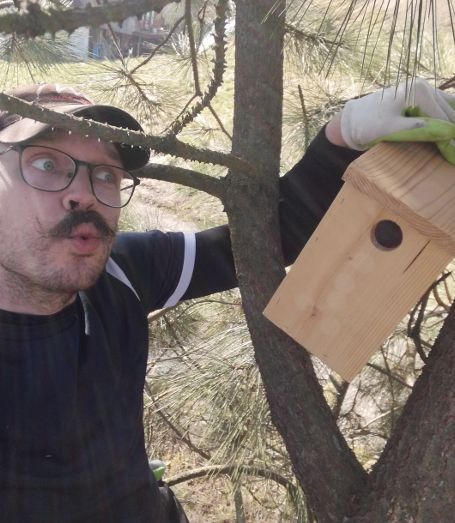 Mężczyzna z wąsami i okularach, w rękawicach, trzyma budkę dla ptaków na tle drzewa iglastego, z wyrazem zdziwienia na twarzy.