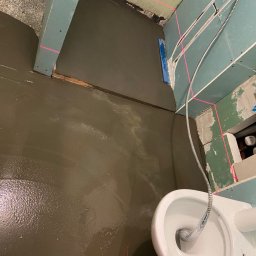 Wiesław Siłka - Wylewka samopoziomująca na podłodze w łazience w trakcie remontu, widoczny sedes, ściany z płyt kartonowo-gipsowych i poziomica laserowa wyznaczająca poziom.