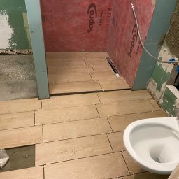 Wiesław Siłka - Układanie podłogi z płytek imitujących drewno w trakcie prac remontowych łazienki, widoczne krzyżyki dystansowe, poziomica i częściowo zamontowana toaleta.