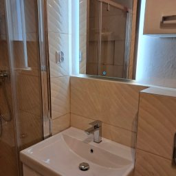 Wiesław Siłka - Biała umywalka z chromowaną baterią i odpływem w nowoczesnej łazience z beżowymi, wzorzystymi płytkami, podświetlanym lustrem i kabiną prysznicową w tle.