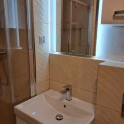 Biała umywalka z chromowaną baterią i odpływem w nowoczesnej łazience z beżowymi, wzorzystymi płytkami, podświetlanym lustrem i kabiną prysznicową w tle.