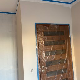 Wiesław Siłka - Przygotowanie do malowania: ściany zabezpieczone taśmą malarską przy suficie i narożnikach, drewniane drzwi oklejone folią ochronną, widoczny włącznik światła.