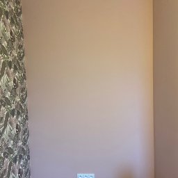 Wiesław Siłka - Ściana pomalowana na pudrowy róż z widocznymi gniazdkami elektrycznymi, fragmentem ściany z tapetą w liście oraz materiałami i narzędziami budowlanymi na podłodze, sugerującymi prace wykończeniowe.