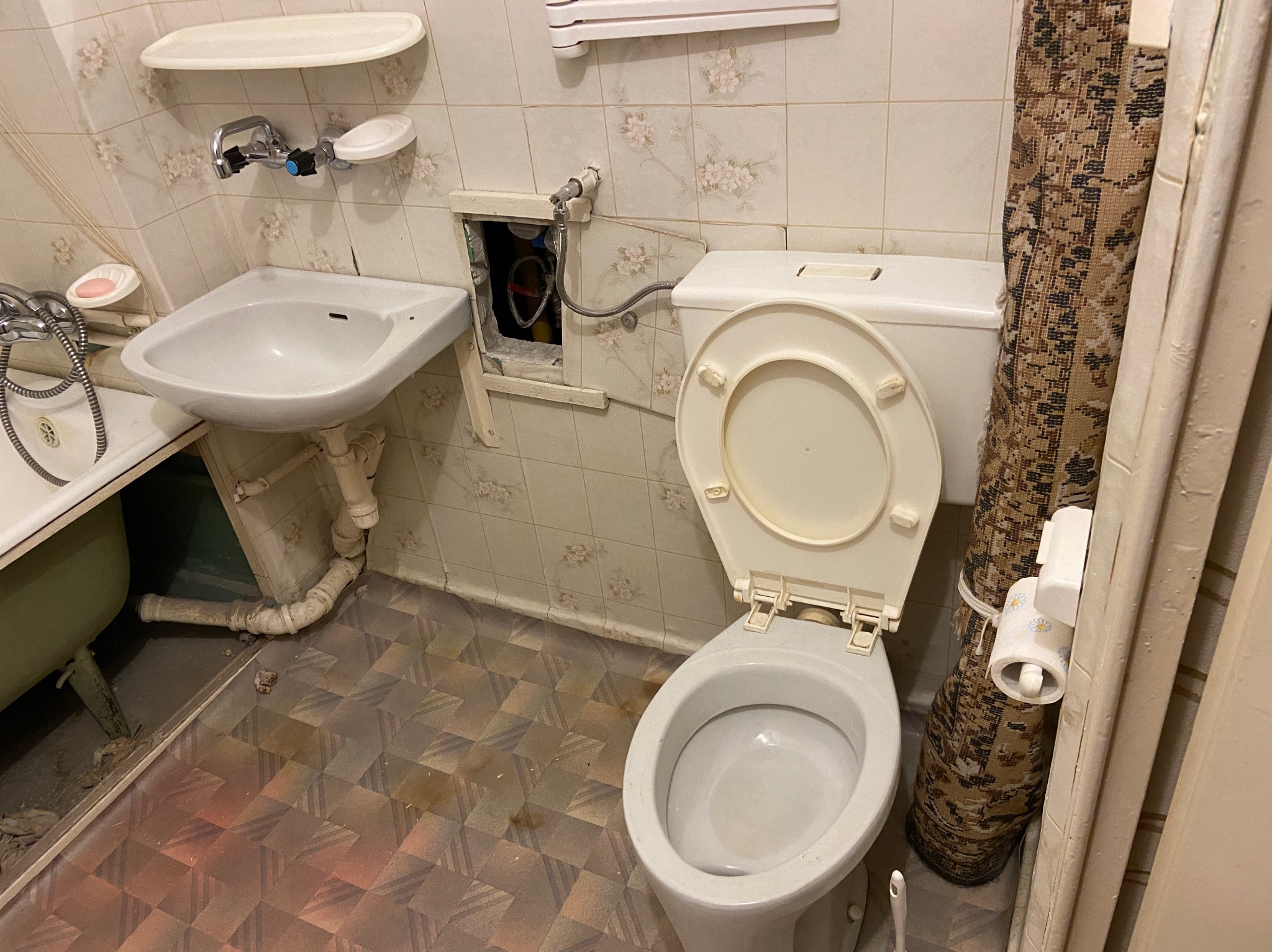 Ujęcie toalety z otwartą klapą, umywalki i wanny w zaniedbanej łazience z płytkami w kwiaty i brudną podłogą, widoczna instalacja hydrauliczna w otworze w ścianie.