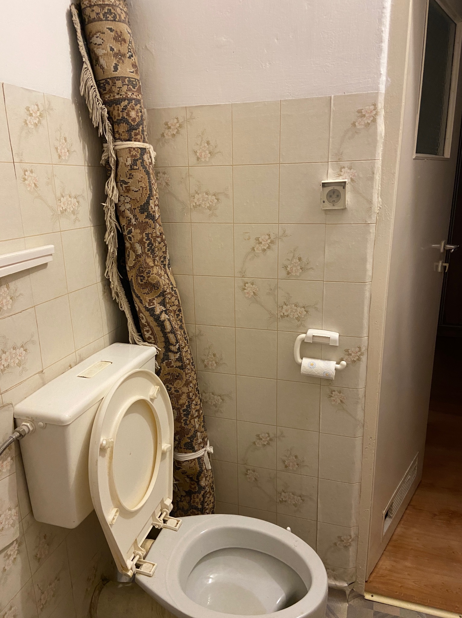 Stara łazienka z beżowymi płytkami w kwiatki, zwinięty dywan oparty o ścianę obok toalety ze zdjętą deską, widoczne ślady użytkowania.