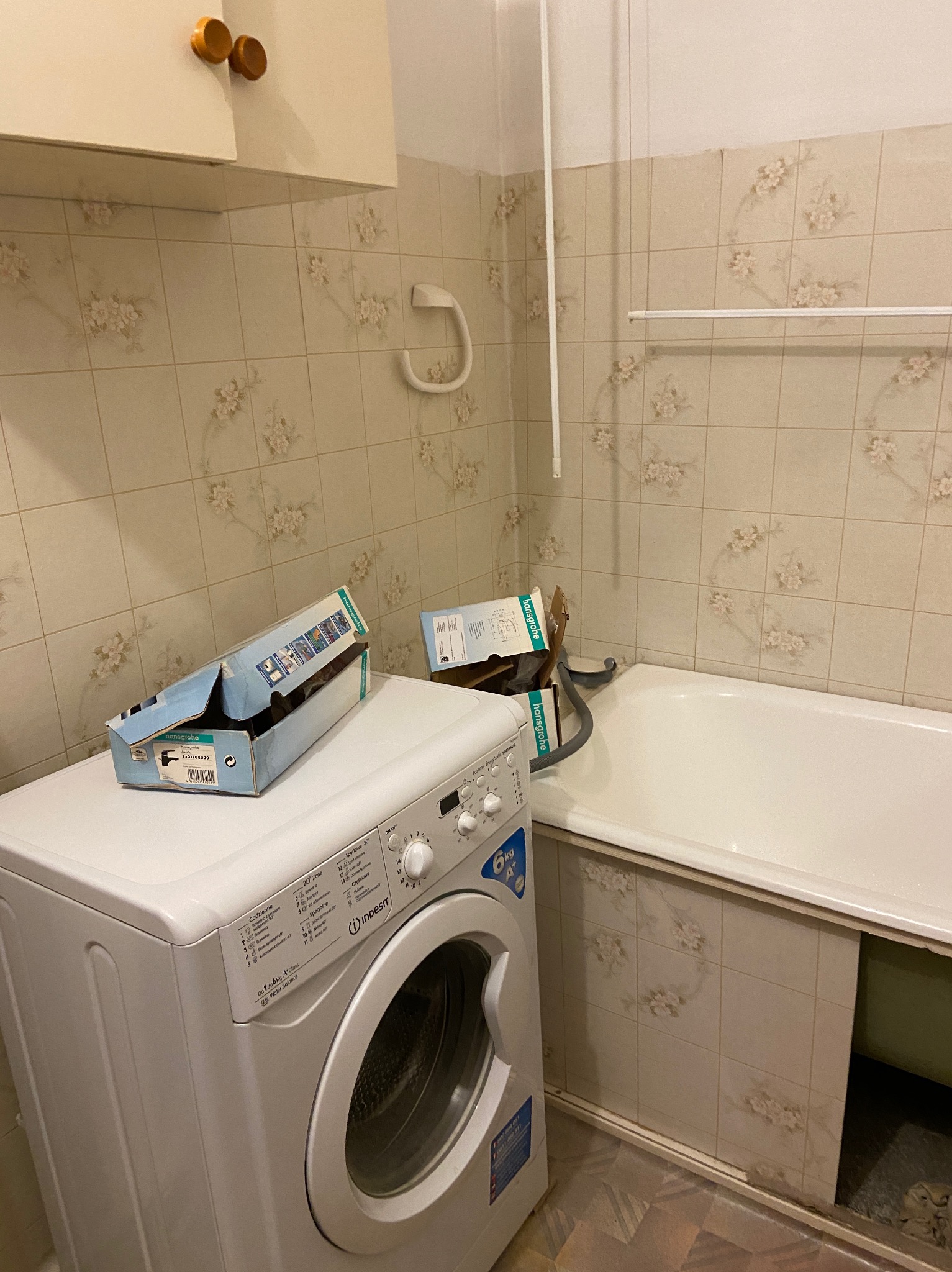 Łazienka z beżowymi płytkami w kwiaty, biała wanna i pralka Indesit z otwartymi kartonami po armaturze Hansgrohe, widoczne akcesoria łazienkowe.