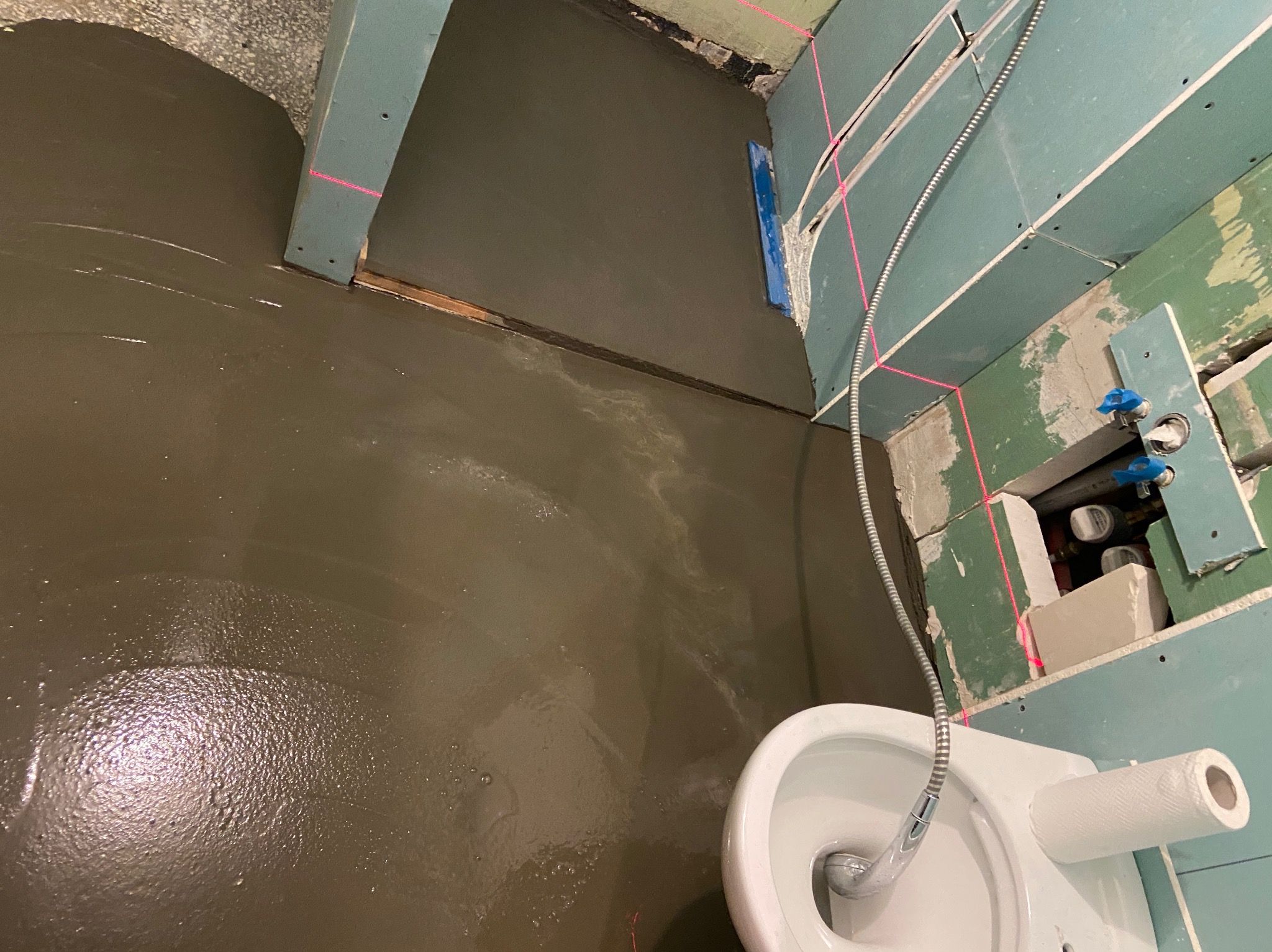 Wylewka samopoziomująca na podłodze w łazience w trakcie remontu, widoczny sedes, ściany z płyt kartonowo-gipsowych i poziomica laserowa wyznaczająca poziom.