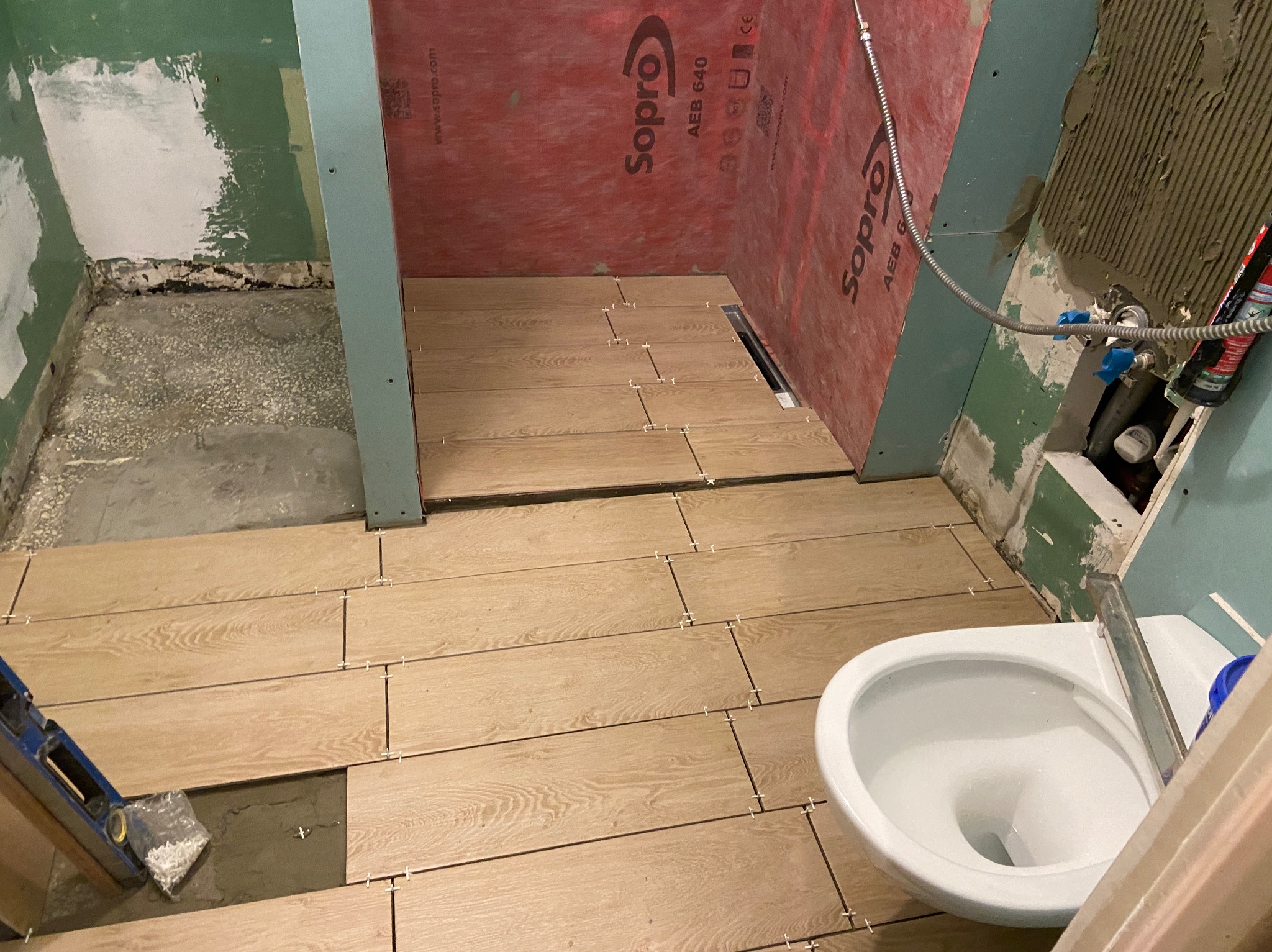 Układanie podłogi z płytek imitujących drewno w trakcie prac remontowych łazienki, widoczne krzyżyki dystansowe, poziomica i częściowo zamontowana toaleta.