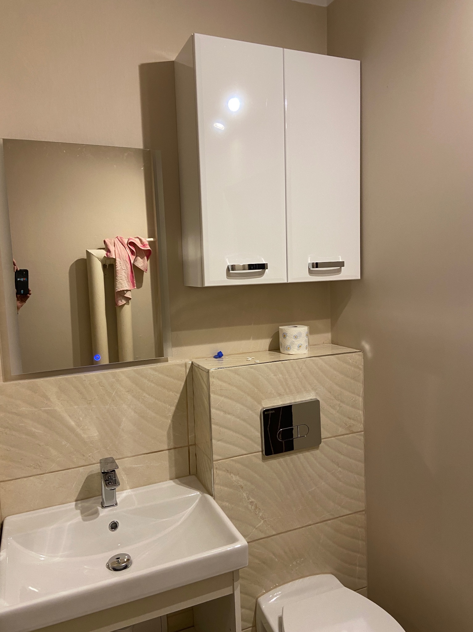 Nowoczesna łazienka z beżowymi płytkami, białym umywalką i szafką nad toaletą, lustro z podświetleniem LED odbija grzejnik i ręcznik.