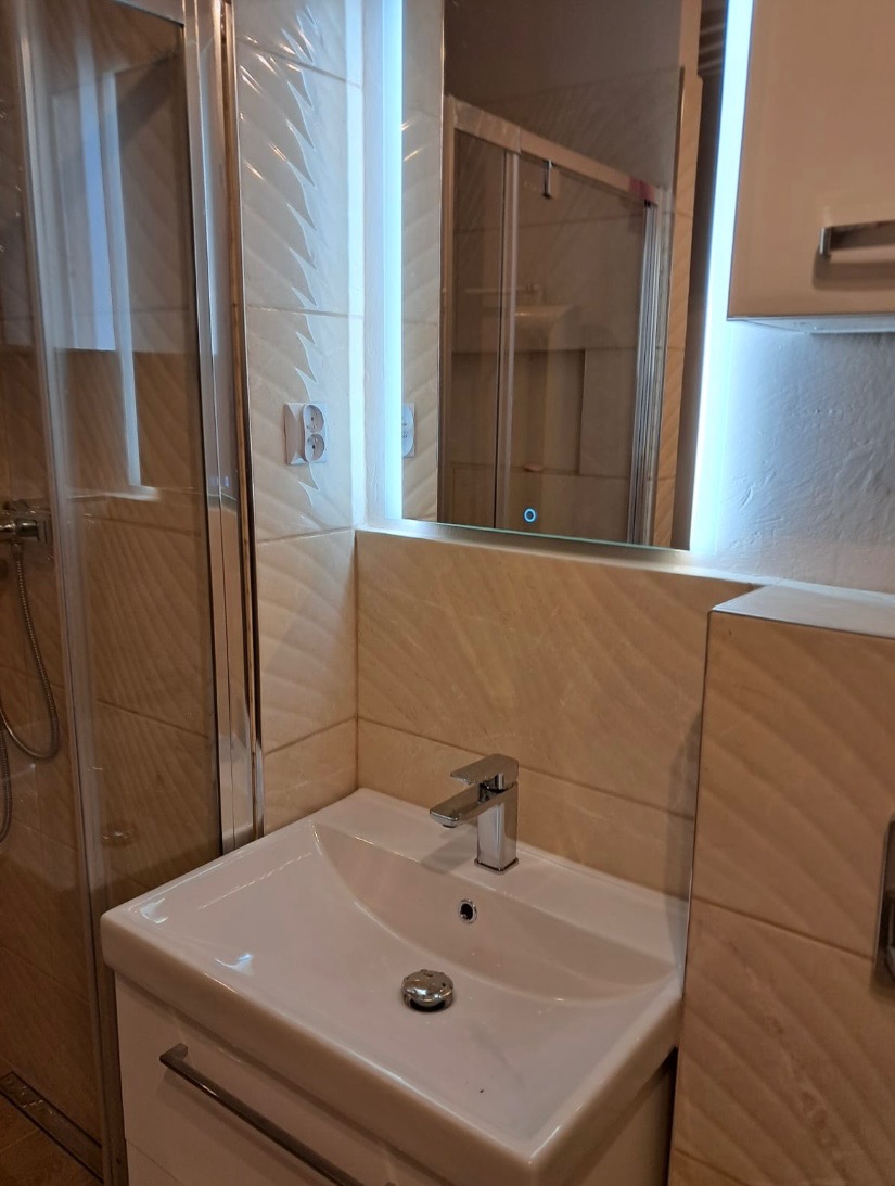 Biała umywalka z chromowaną baterią i odpływem w nowoczesnej łazience z beżowymi, wzorzystymi płytkami, podświetlanym lustrem i kabiną prysznicową w tle.